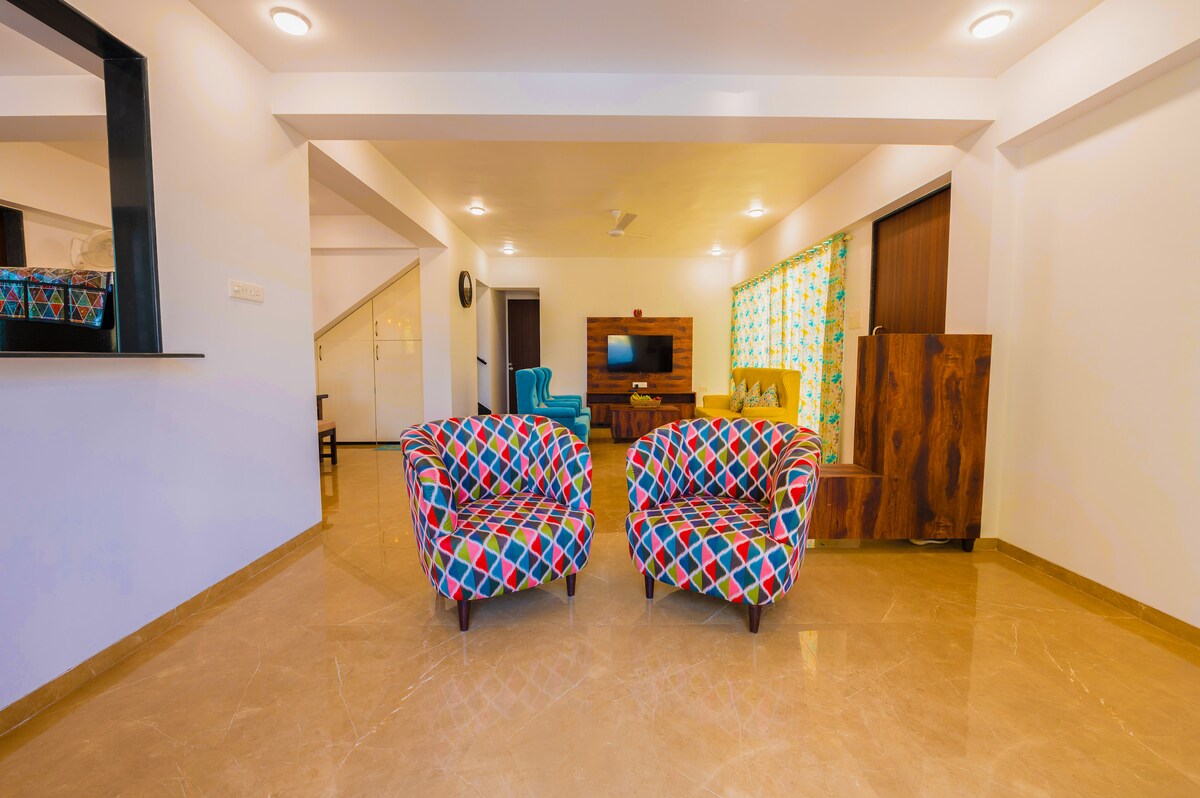 A bright and modern living area is presented, featuring two colorful patterned chairs facing each other. A television is mounted on the wall, complemented by a wooden console. Natural light filters in through the partially covered window with cheerful fabric.