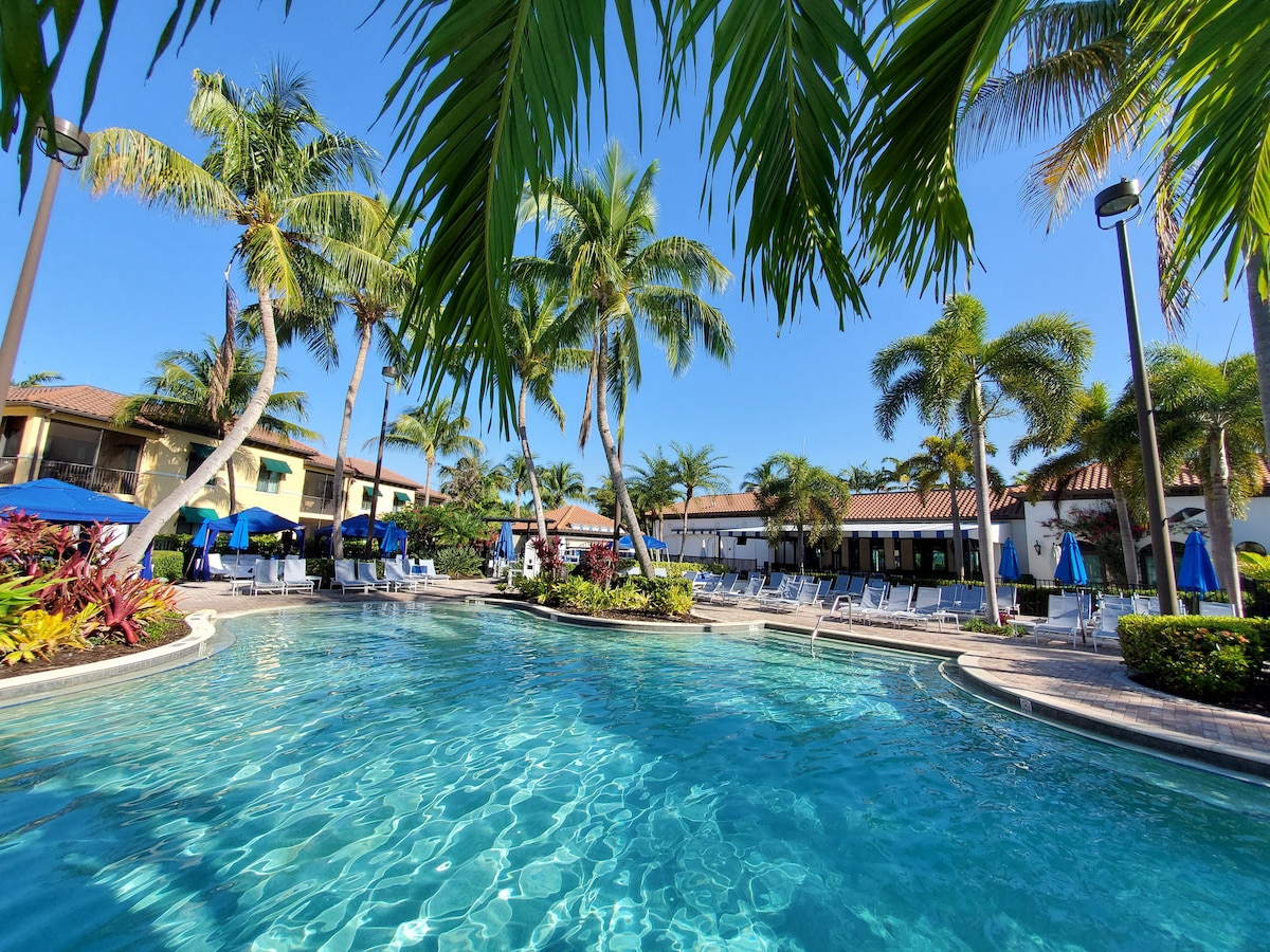 A tranquil pool area is surrounded by lush palm trees and vibrant tropical landscaping. Lounge chairs are placed around the pool, with large umbrellas providing shade. Bright blue skies are visible above, enhancing the inviting atmosphere of the resort-style setting.