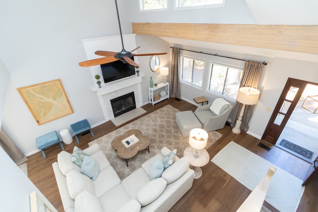 The spacious living room features a cozy sectional sofa and an armchair, arranged around a wooden coffee table. Large windows allow natural light to fill the room, while a fireplace is accentuated by a white mantel. Decorative elements include a wall art piece and floor lamps.