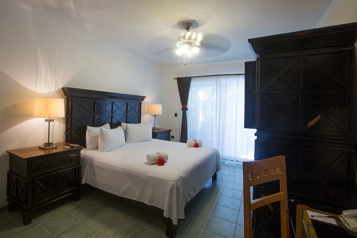 A spacious room features a king-size bed adorned with decorative towels and roses. Two bedside lamps cast a soft glow, while a large cabinet offers ample storage. A small desk is positioned near the window, which is partially draped with dark curtains, allowing light to filter in.