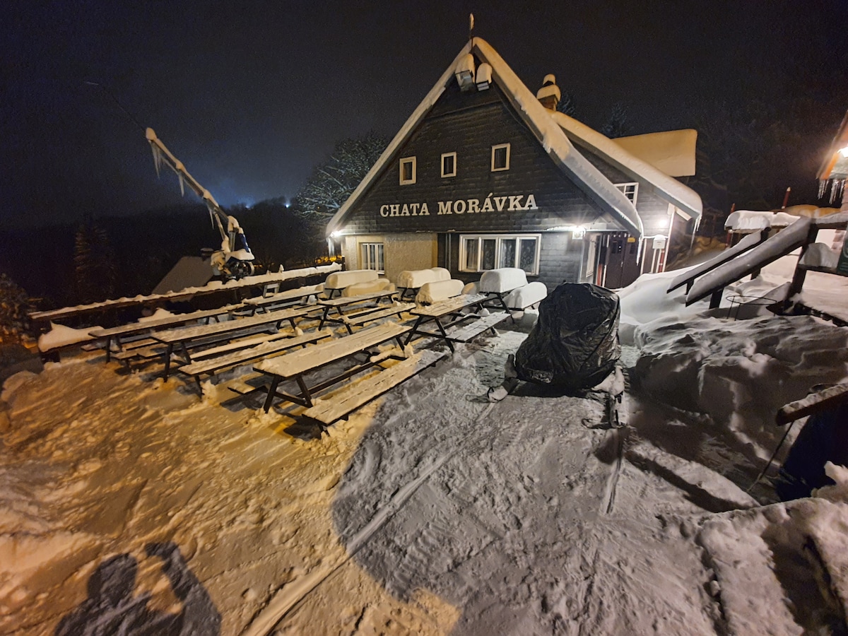 A wooden chalet stands under a night sky, surrounded by snow-covered terrain. Outdoor seating, including multiple benches and tables, is partially buried in snow. Soft lighting from the building highlights the structure, while ski lifts can be seen in the distance amidst a misty atmosphere.