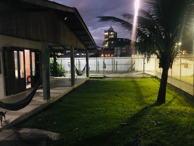 An outdoor area features a well-maintained grass lawn, complemented by two hammocks hanging under a covered porch. Soft lighting illuminates the space, creating a serene environment. The background includes a fence and glimpses of nearby buildings, enhancing the sense of privacy.
