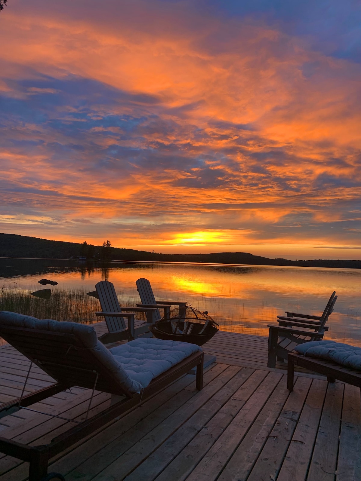 A serene lakeside scene captured at sunset, with vibrant orange and purple hues reflected in the calm water. Comfortable lounge chairs are positioned on a wooden deck, providing a perfect spot for relaxation while taking in the stunning views.