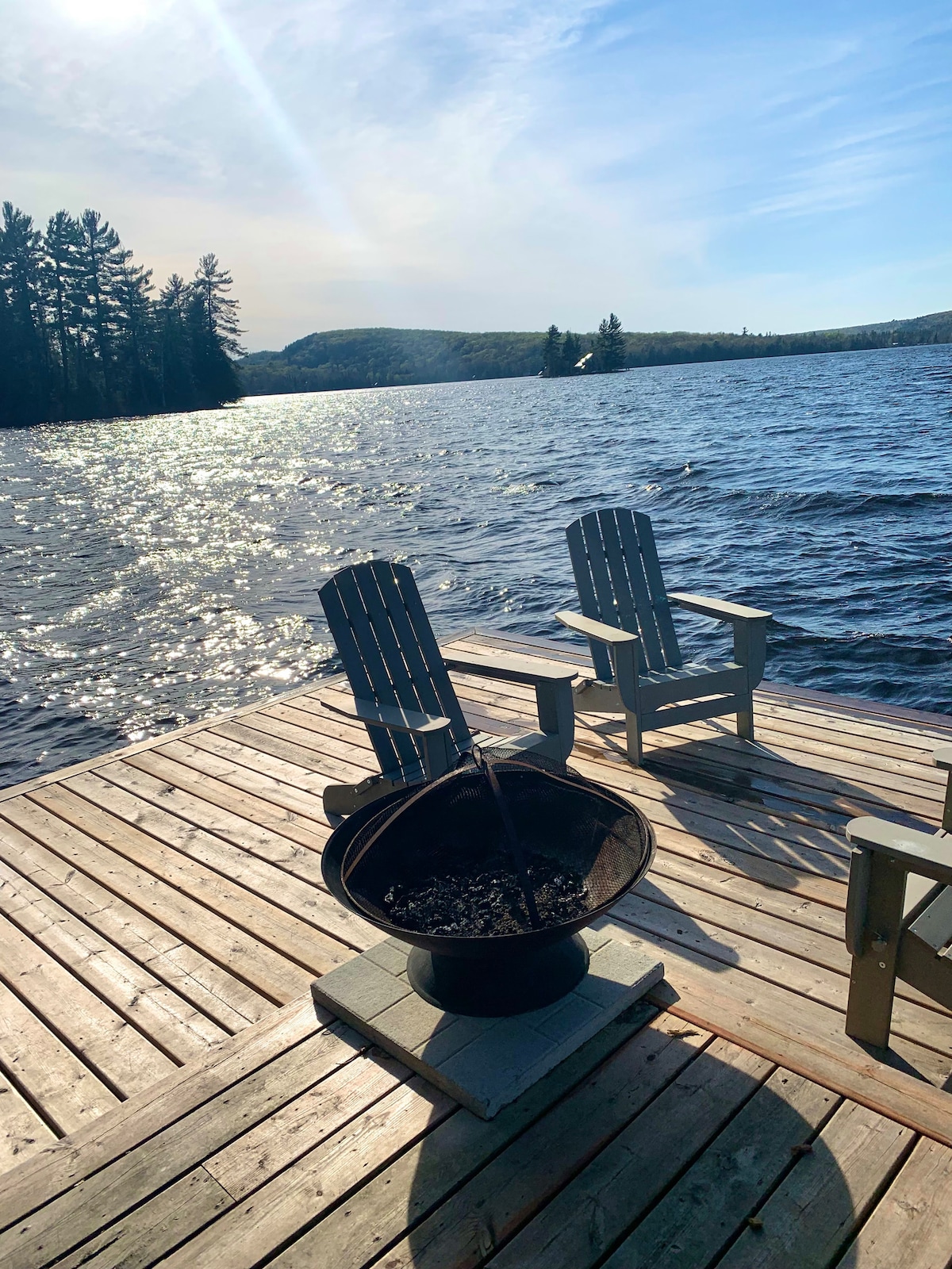 A wooden dock extends over the shimmering water, with comfortable Adirondack chairs positioned nearby. A fire pit sits at the center, inviting gatherings. Tall trees bordering the lake reflect in the water, while rolling hills are visible in the distance under a clear blue sky.