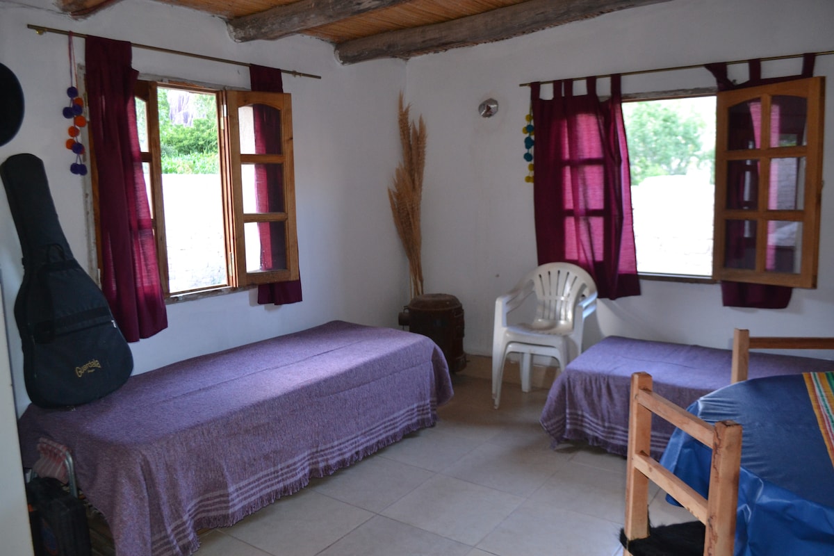 A cozy interior space features two single beds draped in purple blankets. Light streams through two open wooden windows adorned with dark curtains. A white chair is positioned beside a simple wooden table, enhancing the room's functionality, while a guitar rests against the wall.