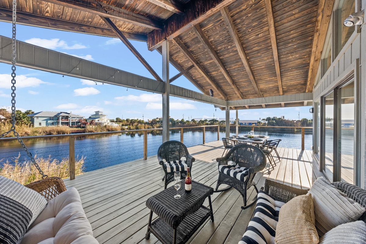 A spacious wrap-around porch deck provides a serene view of the lake. Comfortable seating, including a cushioned couch and woven chairs, is arranged around a coffee table. The open structure features a wooden ceiling with beams, allowing natural light to enhance the relaxing atmosphere.