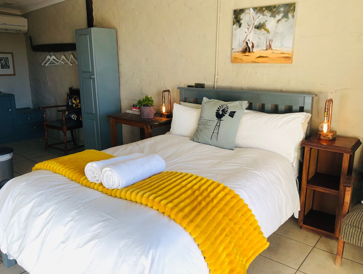 A spacious bed is dressed in crisp white linens, accented by a bright yellow throw. Wooden bedside tables with lamps flank the bed. An art piece hangs above, featuring a serene landscape. Additional furniture includes a blue cabinet and a chair in the background.