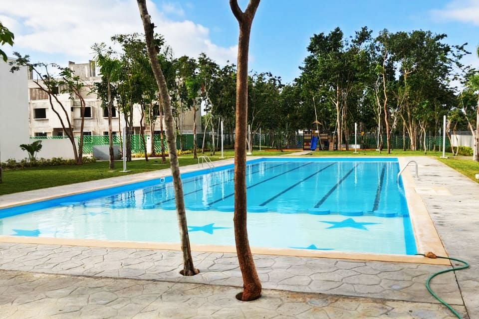 A semi-Olympic pool is surrounded by well-maintained green spaces and trees. Bright blue water reflects the clear sky, while the poolside area features a smooth stone pathway. Additional recreational facilities are visible in the distance, contributing to a relaxing outdoor environment.