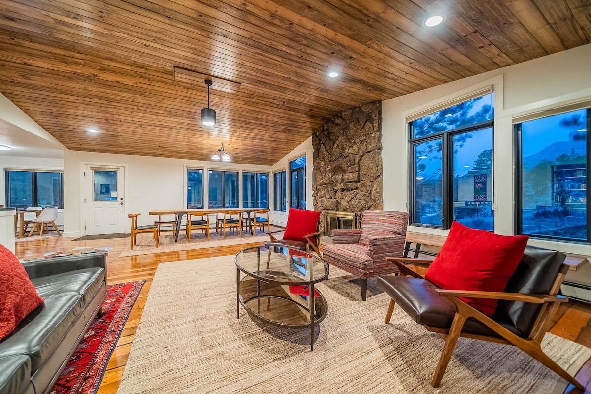 A spacious living area features hardwood floors and a warm wooden ceiling. Large windows provide views of the surrounding landscape. Comfortable seating includes a leather sofa with red cushions and two accent chairs. A glass coffee table centers the room, complemented by a colorful area rug.