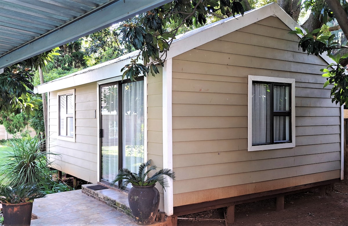 The exterior of the cottage is showcased, featuring a light-colored wooden structure with large glass doors and a nearby window. Potted plants are positioned at the entrance, and a shaded area is provided by a roof overhang. Lush greenery is visible in the background.