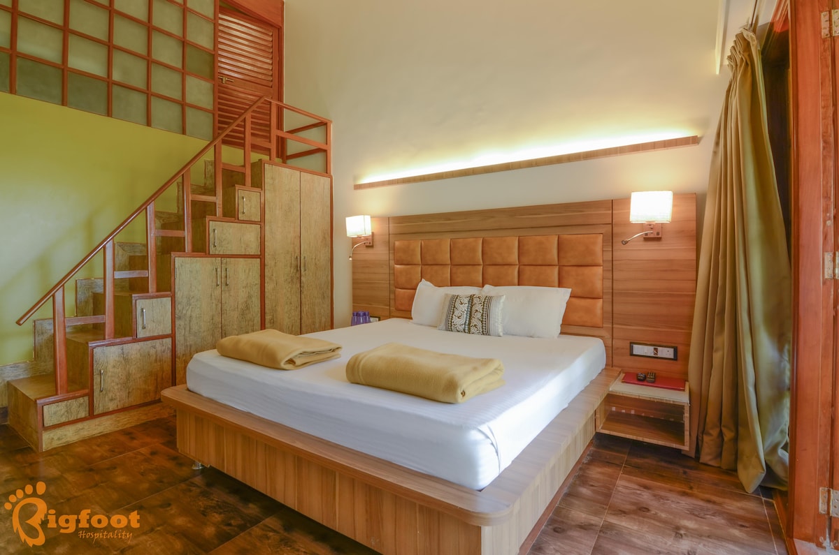 A spacious bedroom features a king-size bed with neutral bedding and two golden towels folded on top. The room is complemented by wooden accents and soft lighting, with a staircase leading to an upper area. Warm green walls add a subtle contrast to the wooden furnishings.