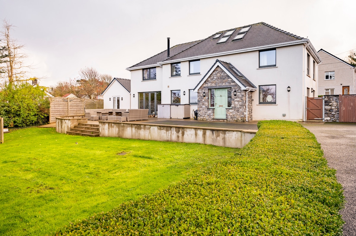 An expansive view of the farmhouse showcases a modern exterior with a combination of stone and white siding. The landscaped garden features a manicured lawn bordered by shrubs, with a large stone patio area accessible from the house.