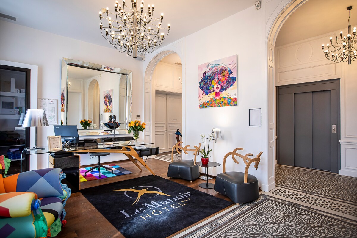 The lobby area features an elegant chandelier and a stylish reception desk with a large mirror. Colorful seating is provided, accompanied by artworks on the wall. An elevator is visible, and a patterned rug enhances the welcoming atmosphere of the space.