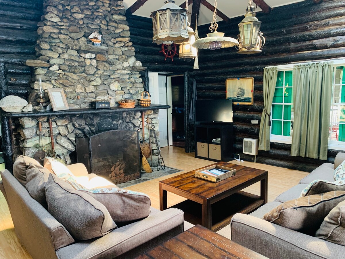 A spacious living area showcases a stone fireplace as the focal point, surrounded by comfortable seating arrangements. Lantern light fixtures hang from the ceiling. A television is positioned near the entrance, with green curtains framing the windows, allowing natural light to enhance the rustic ambiance.