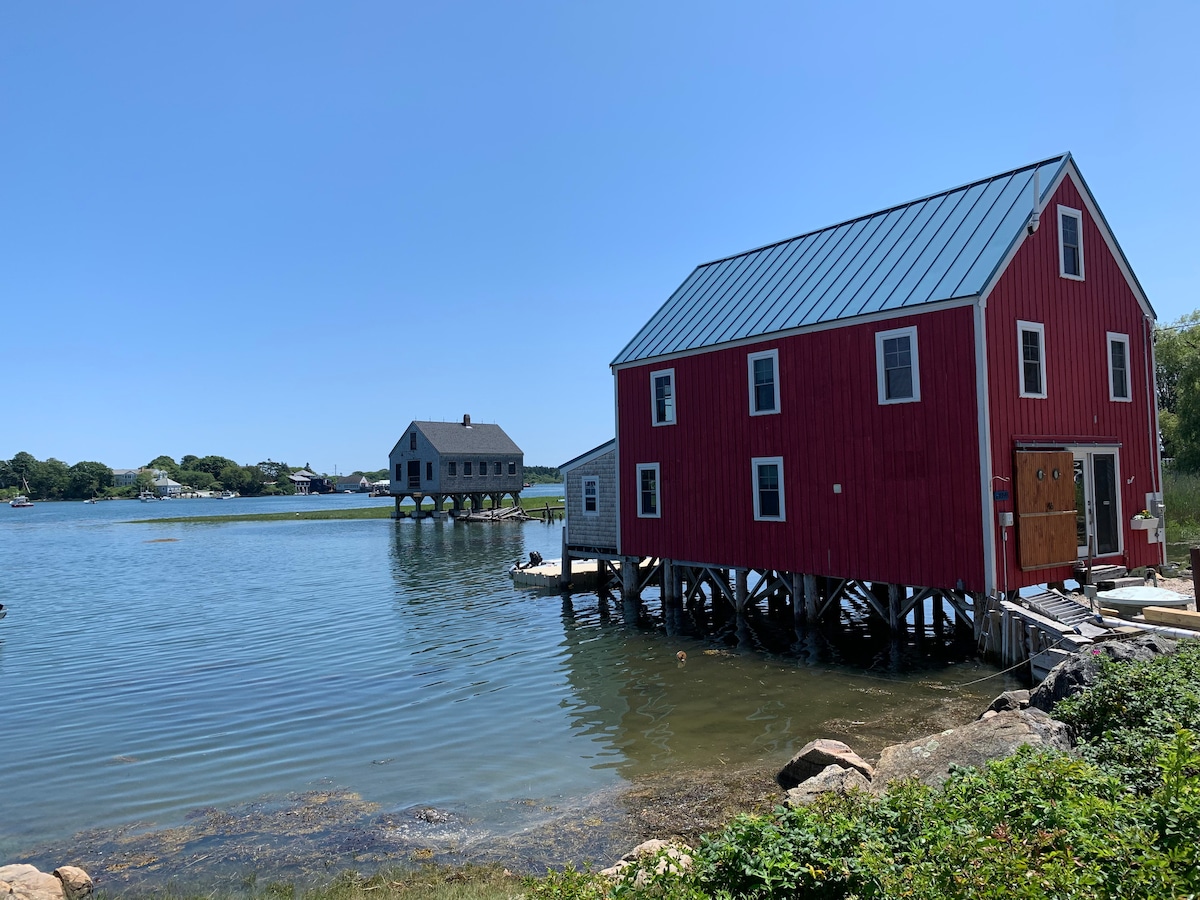 A vibrant red building on stilts is positioned along a calm body of water, reflecting the clear blue sky above. In the background, a darker building sits across the water, contributing to a serene coastal scene.