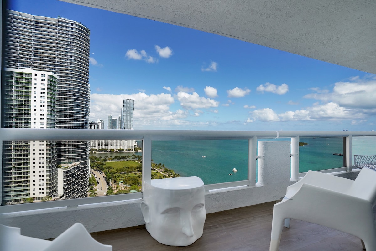 The private balcony offers sweeping views of Biscayne Bay and Margaret Pace Park, with modern white seating providing a comfortable space to relax. The surrounding high-rise buildings frame the scene, while the clear blue sky enhances the serene atmosphere.