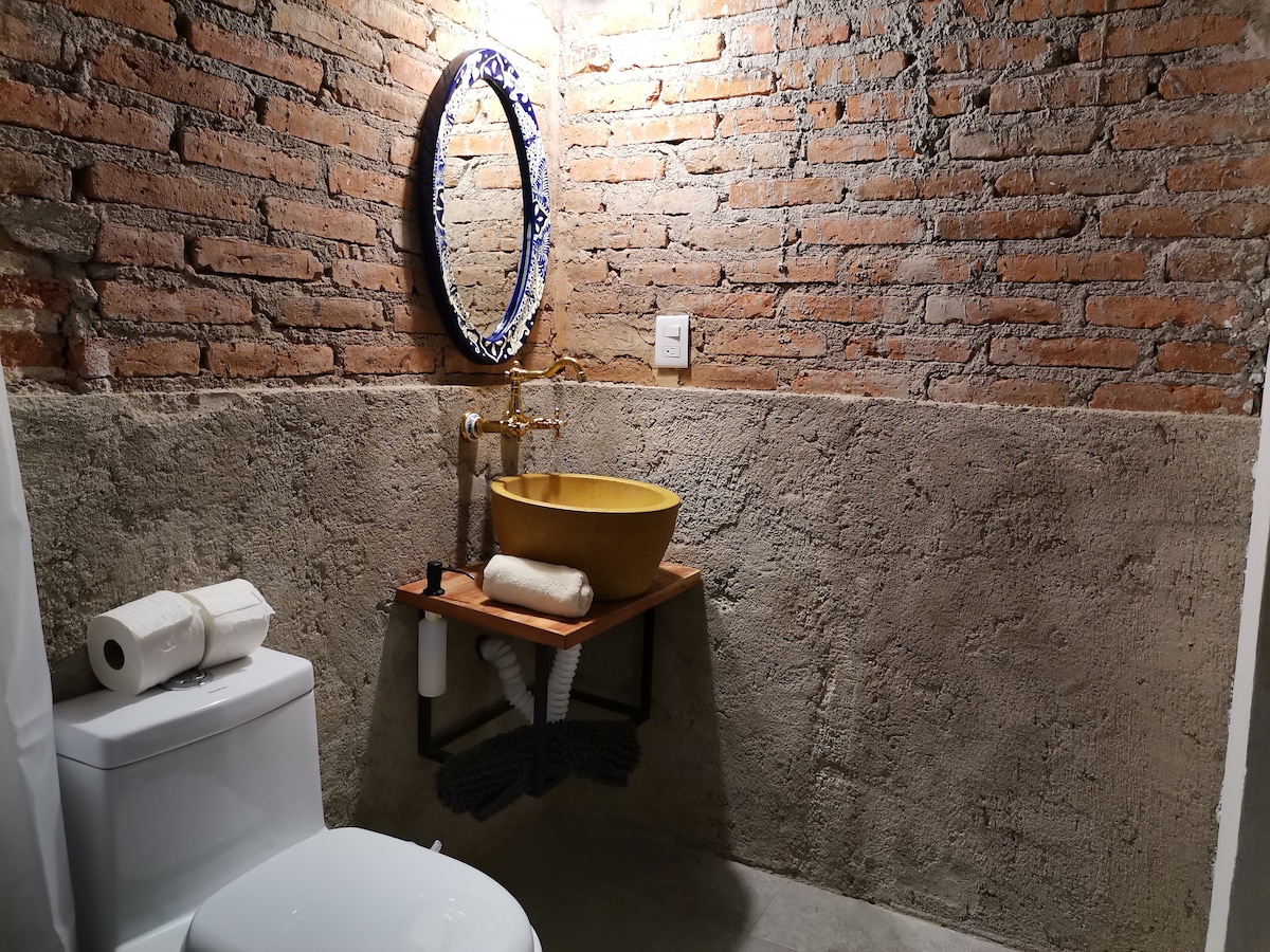 A modern bathroom features exposed brick walls and a concrete finishing. A golden bowl sink sits atop a wooden shelf, with elegant gold hardware. A toilet is positioned adjacent, and essential toiletries are neatly arranged beside the sink. Soft lighting enhances the unique textures of the space.