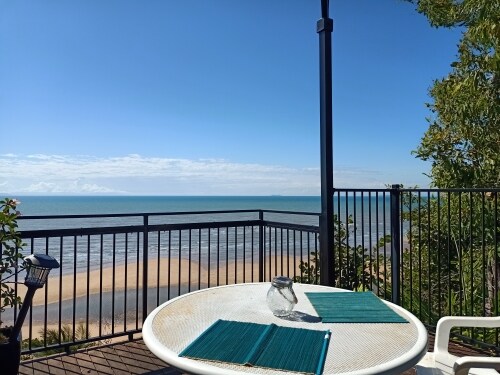 A round outdoor table is set on a balcony, offering views of the beach and the sea. Two teal placemats lie on the table, with a glass jar placed in the center. A railing surrounds the deck, and nearby greenery adds a natural touch.