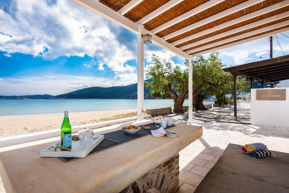 An outdoor dining area is presented under a wood-beamed structure, overlooking a sandy beach and serene blue waters. A stone table holds a tray with beverages and snacks. Lush greenery and distant mountains are visible, creating a peaceful coastal environment.