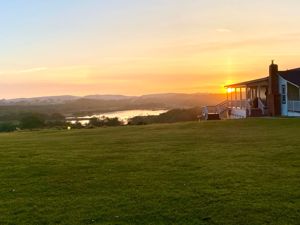 A serene sunset view from the expansive green lawn, showcasing the peaceful reflection of the orange and yellow hues in the calm water. The cottage, with a spacious porch, stands to the right against a backdrop of rolling hills.