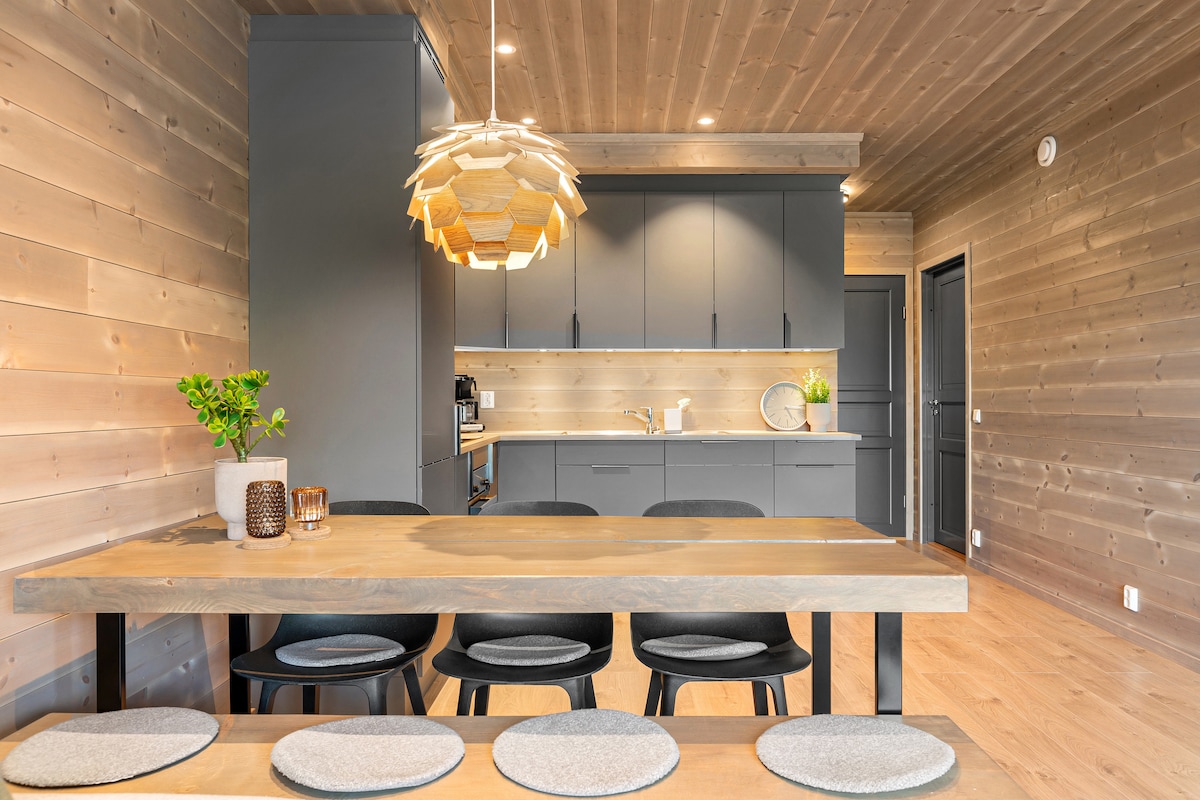 A modern dining area features a long wooden table surrounded by several upholstered chairs. A pendant light hangs above the table, illuminating the space. The kitchen area, with a sleek design and wooden accents, is visible in the background.
