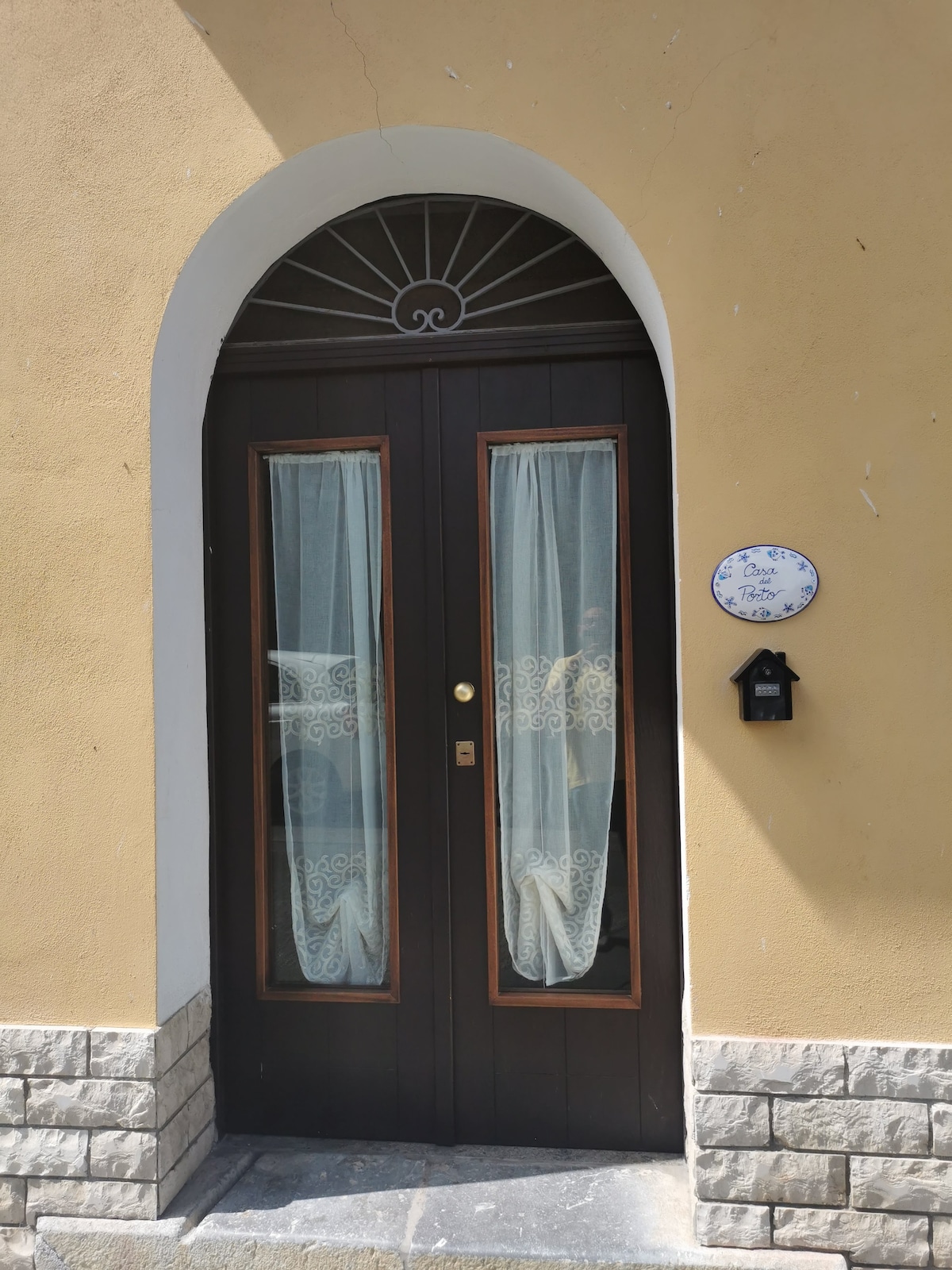 A traditional entrance is displayed, featuring a wooden door with elegant arched windows adorned with light, sheer curtains. The stonework at the base adds a rustic touch, while a decorative house number plaque is positioned beside the door.