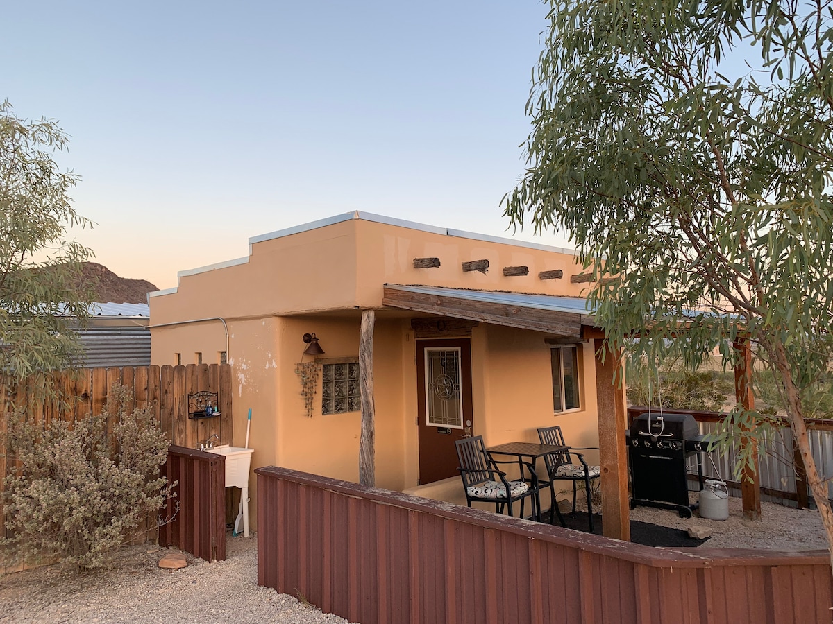 The exterior of a casita is showcased, featuring warm earth-toned walls and a modern flat roof. Outdoor seating with metal chairs is visible on a small patio area, accompanied by a propane grill. Native shrubs and landscapes surround the structure, providing a natural desert ambiance.
