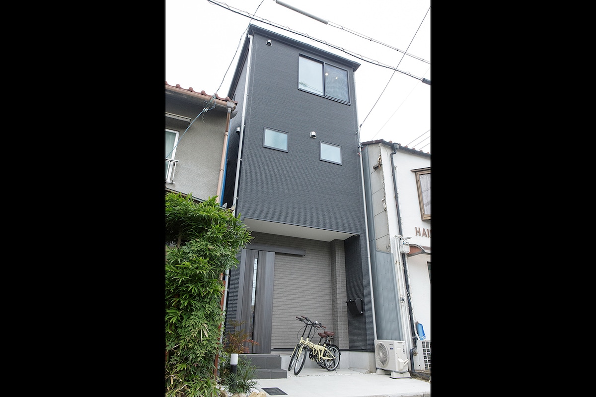 A modern three-story building is depicted, featuring a sleek exterior with dark cladding and multiple windows. A small paved entrance is visible, with two bicycles parked outside, surrounded by well-kept greenery.