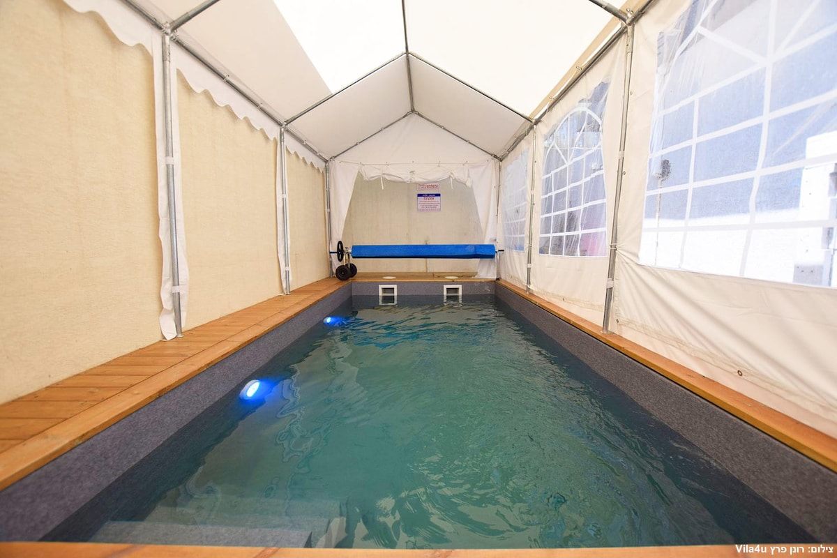 A covered pool area features a rectangular pool with clear water, illuminated by subtle blue lights. Surrounding the pool is a wooden deck, and the tent walls are made of fabric with large windows, providing a sense of openness while maintaining protection from the elements.