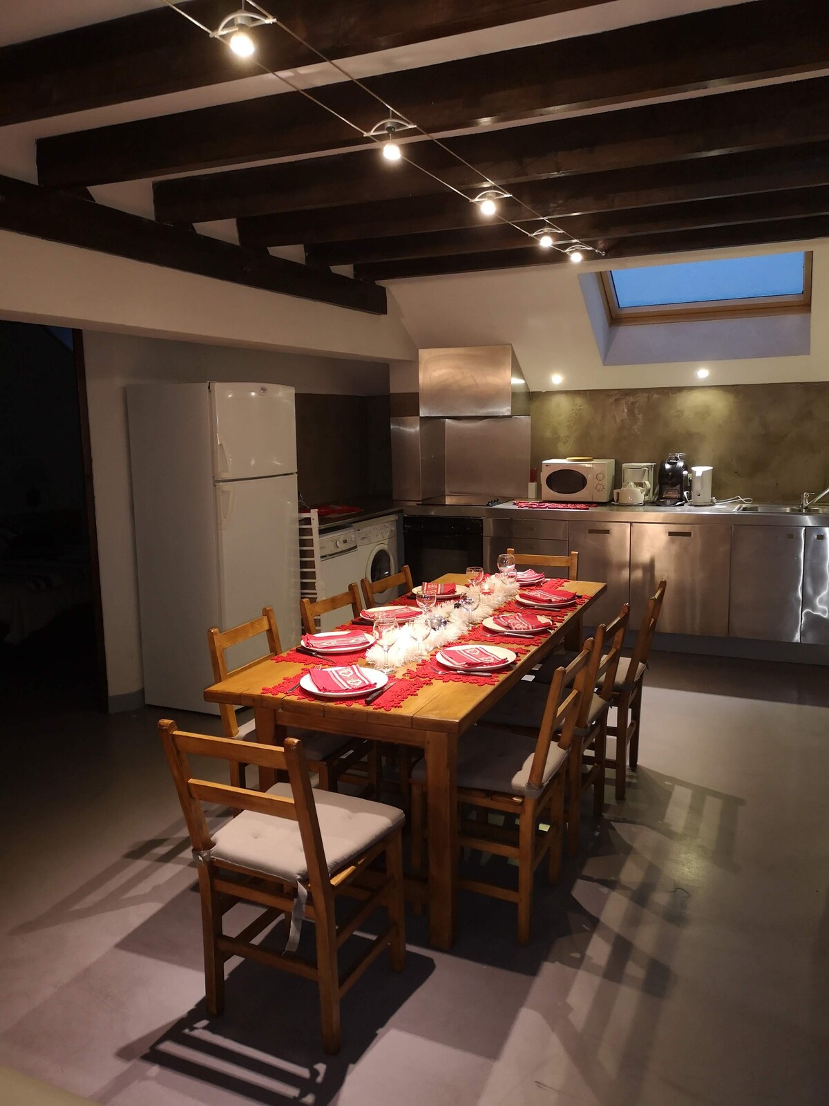 A spacious dining area is presented with a wooden table set for six, featuring red tableware. The kitchen is equipped with modern appliances, including a refrigerator and an oven. Soft overhead lighting creates a warm ambiance, complemented by a skylight allowing natural light to enter.