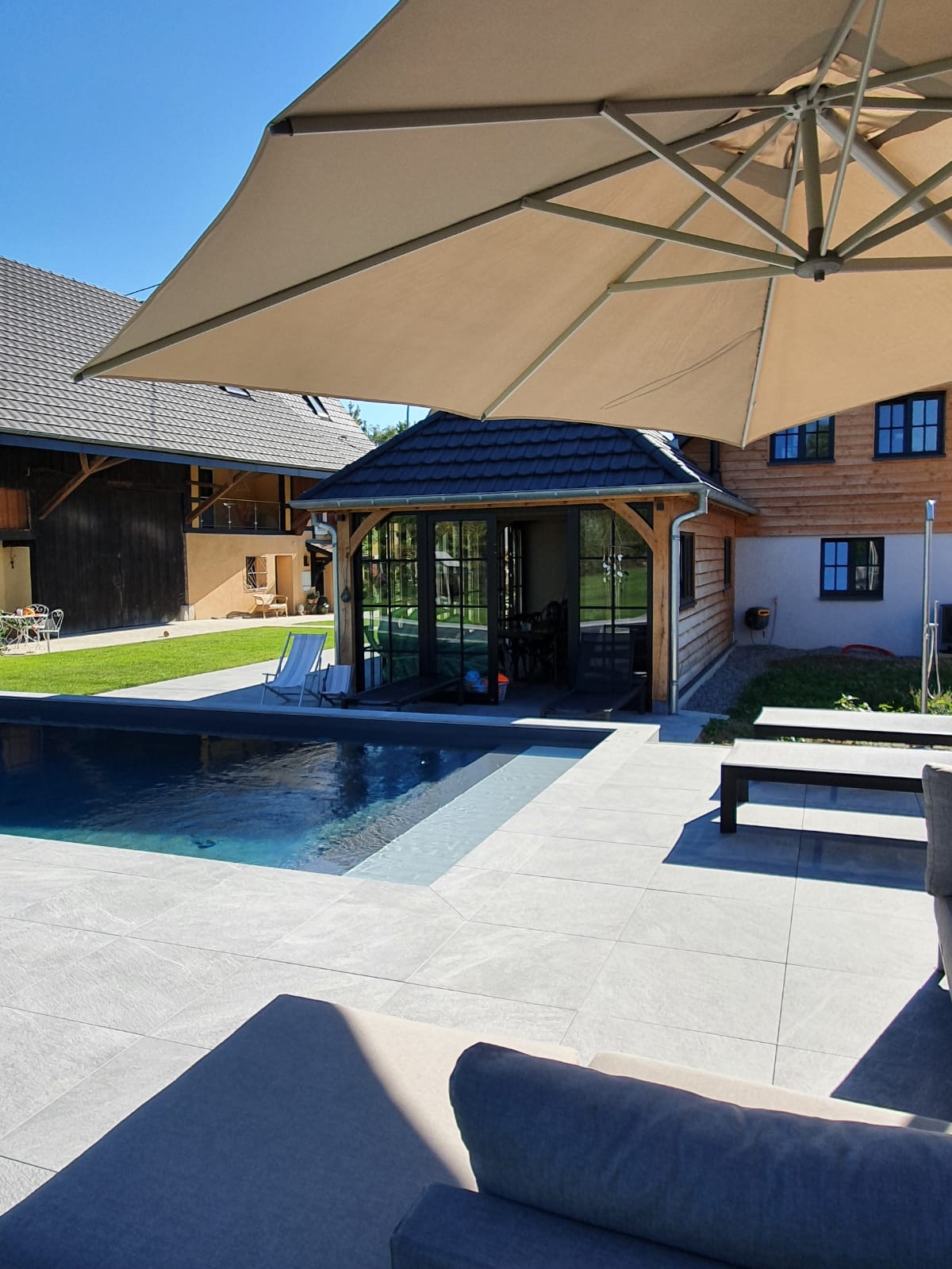 A view of the outdoor area features a swimming pool surrounded by sleek stone tiles. A shaded seating area is visible, with an umbrella providing respite from the sun. Green grass frames the pool, enhancing the serene environment.