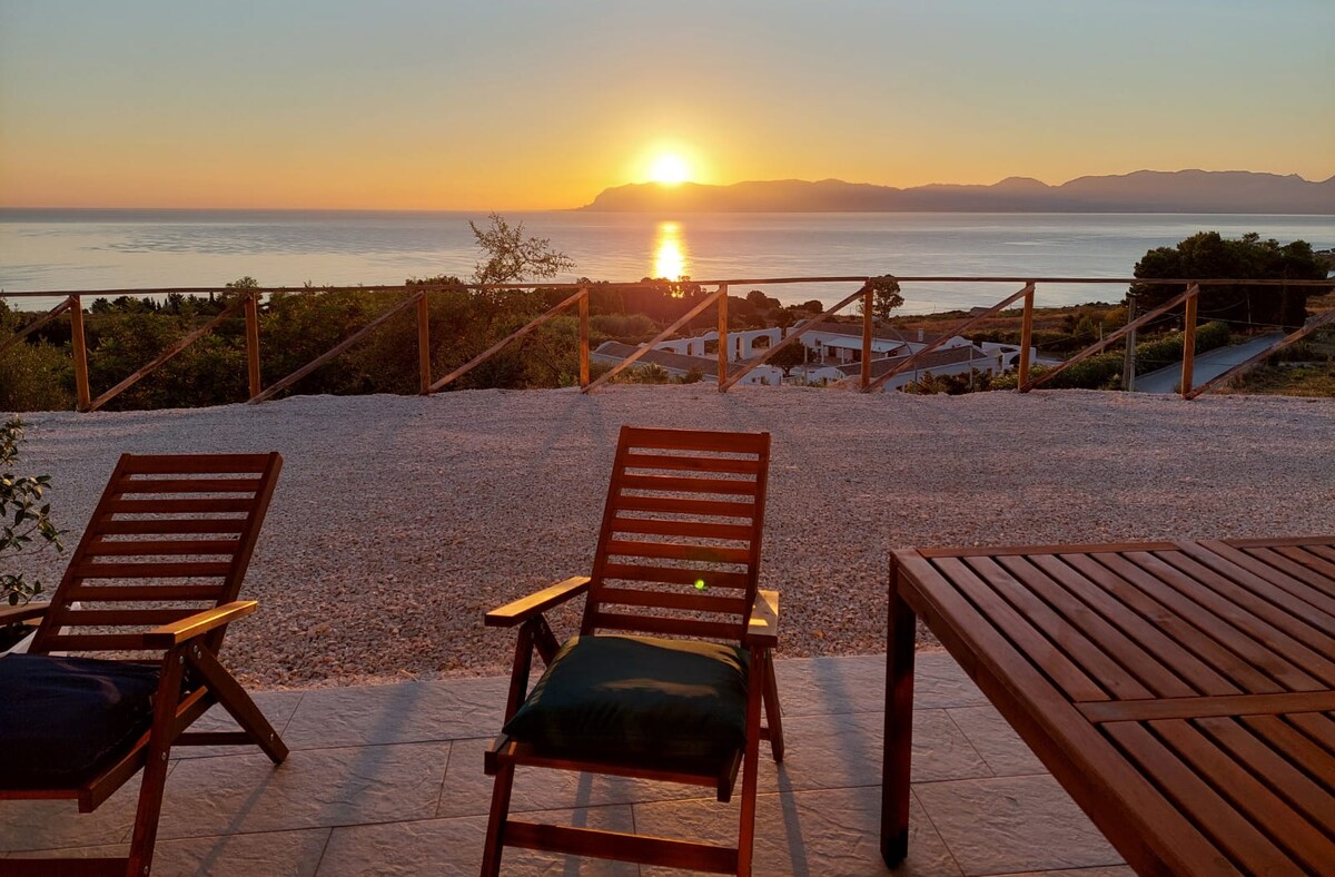 The image captures a tranquil sunset over the sea, viewed from a patio with two wooden lounge chairs. A wooden table complements the seating area, set against a backdrop of sweeping coastal views and the distant silhouette of hills.