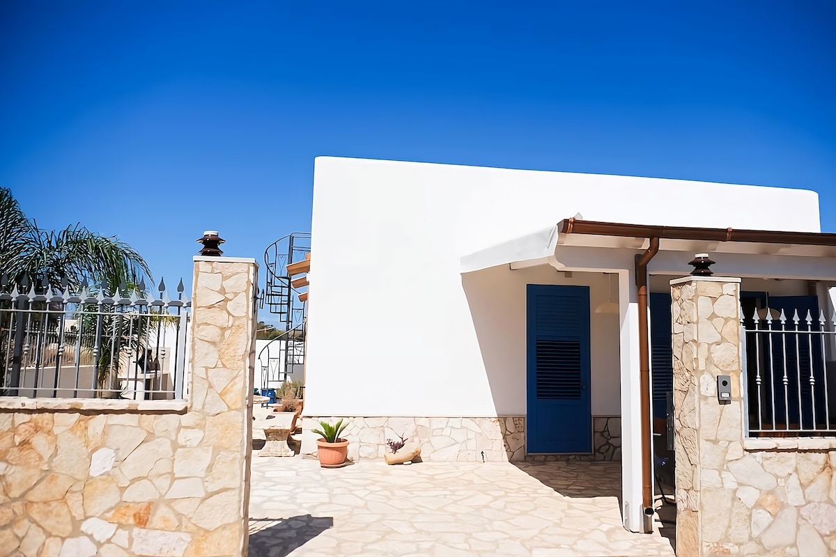 The exterior view of a white-walled structure features a stone pathway leading to an entrance. A blue door adds a pop of color, while a spiral staircase can be seen in the background. Lush greenery enhances the welcoming atmosphere.