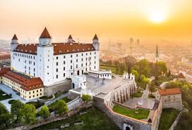 The aerial view captures a historic castle with distinctive red rooftops and four prominent towers, situated on a hillside. Lush greenery surrounds the structure, and the scene is bathed in warm golden light from the setting sun, with a cityscape visible in the background.