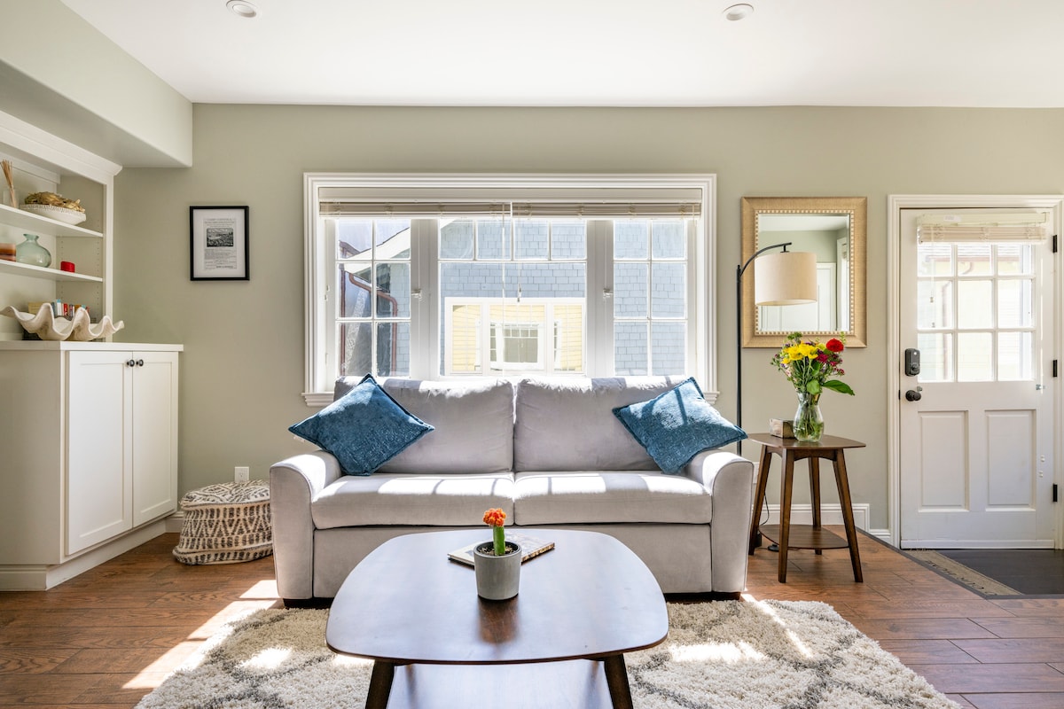 A comfortable living area features a light gray sofa adorned with blue cushions, situated next to a wooden coffee table with a small plant. Natural light floods in through large windows, illuminating the space. A mirror and a vase of flowers are displayed on adjacent surfaces.