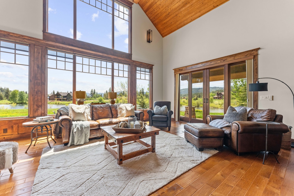 A spacious living area features large floor-to-ceiling windows that allow natural light to fill the room. Comfortable leather seating is arranged around a wooden coffee table, while a cozy area rug lies beneath. Views of the surrounding greenery and private pond can be seen through the windows.