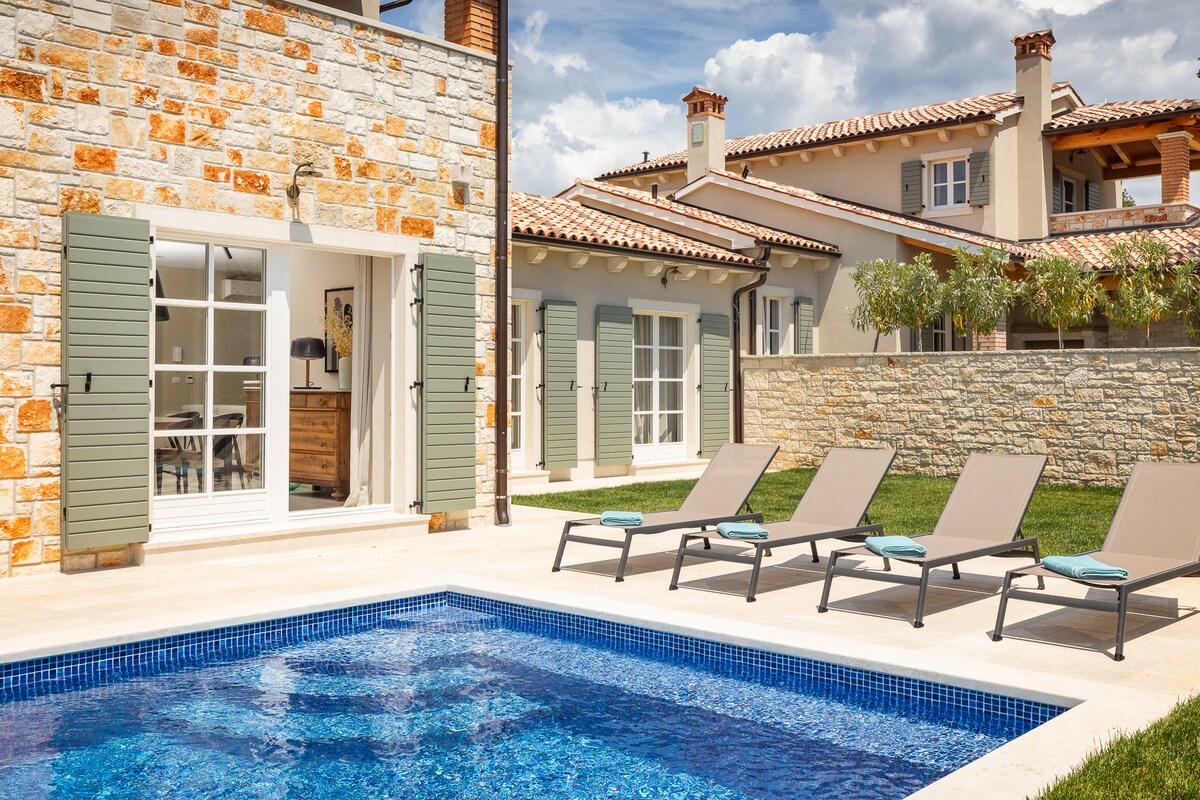 The image features a private swimming pool surrounded by sun loungers, located in a well-maintained outdoor area. The villa's stone exterior and green shutters are visible, along with a spacious terrace area. Lush grass borders the pool, providing a relaxing environment.