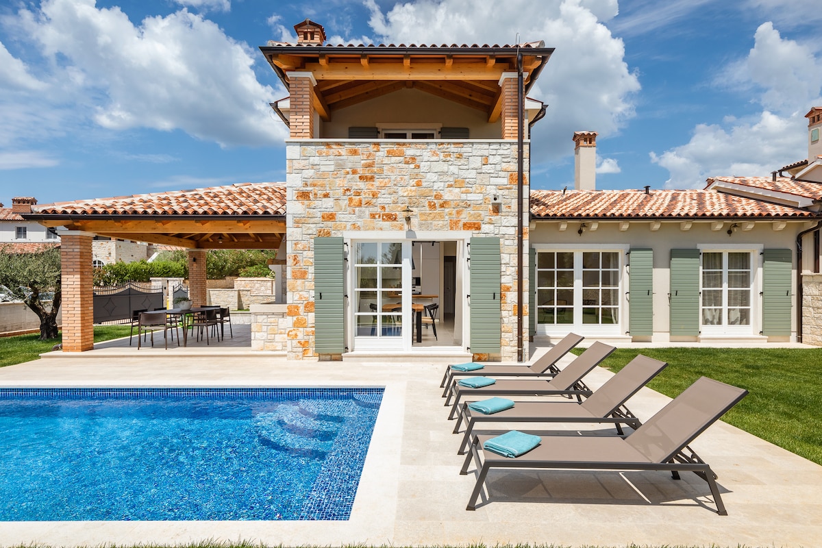 The exterior of the villa showcases a private swimming pool surrounded by lounges. Stone and wood architecture is complemented by green window shutters. A spacious terrace is visible, with a dining area and landscaped garden, enhancing the outdoor living experience.