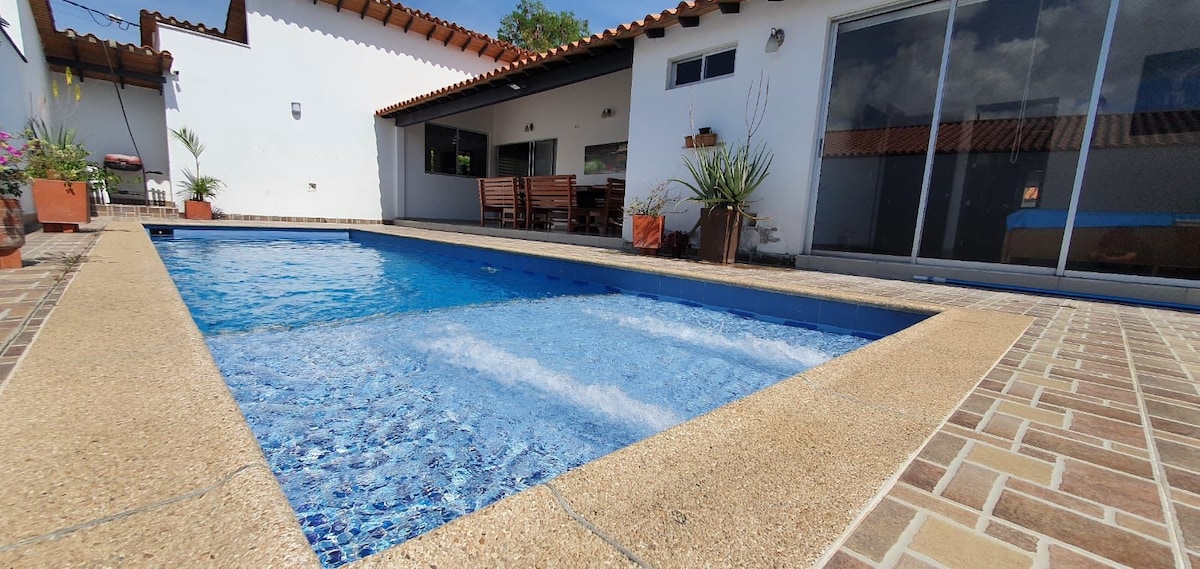 A swimming pool with clear blue water occupies the center of the outdoor area, bordered by textured stone tiles. Wooden lounge chairs are placed nearby, accompanied by potted plants. The house's exterior features large windows reflecting natural light and providing views of the surrounding landscape.