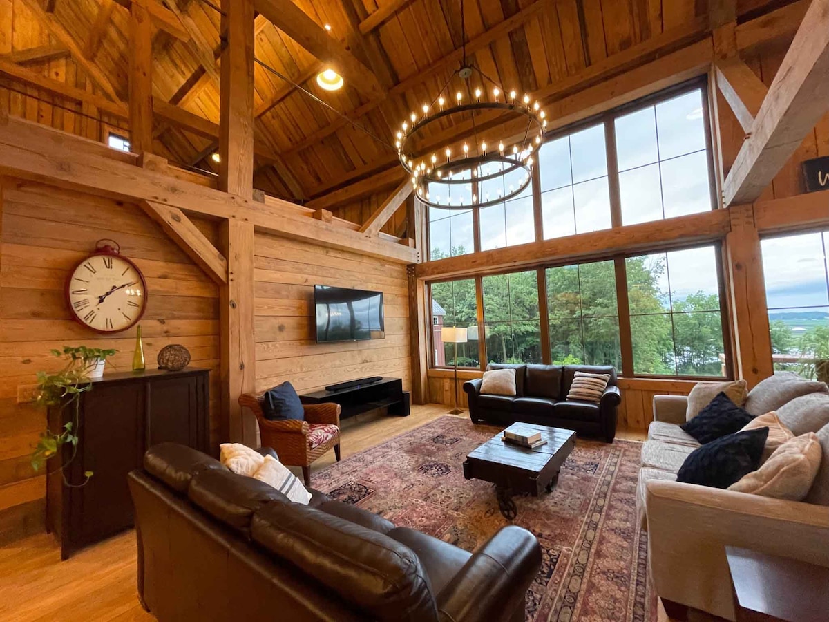 A spacious living area features high wooden ceilings supported by exposed beams. Large windows allow natural light to fill the room, highlighting a cozy seating arrangement with leather and upholstered sofas. A chandelier hangs above a central coffee table, contributing to the warm ambiance.