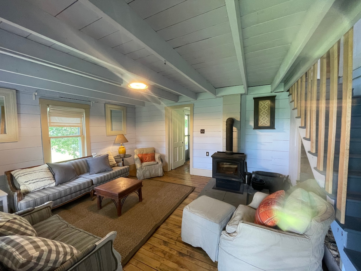 A rustic living area is presented with comfortable seating options, including plush sofas and a patterned area rug. A wooden coffee table sits centrally, with natural light filtering through two windows. A wood-burning stove enhances the cozy feel, complemented by original wood beams and flooring.