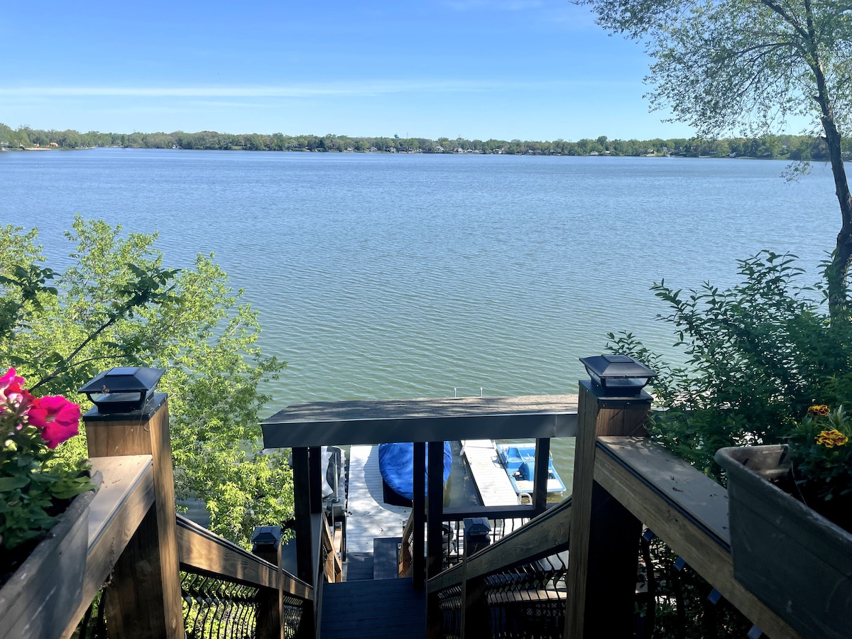 A scenic view of the lake is captured from a wooden staircase lined with greenery and flowers. The calm water reflects a clear blue sky, and a private pier is visible at the water's edge, ready for relaxed lakefront activities.