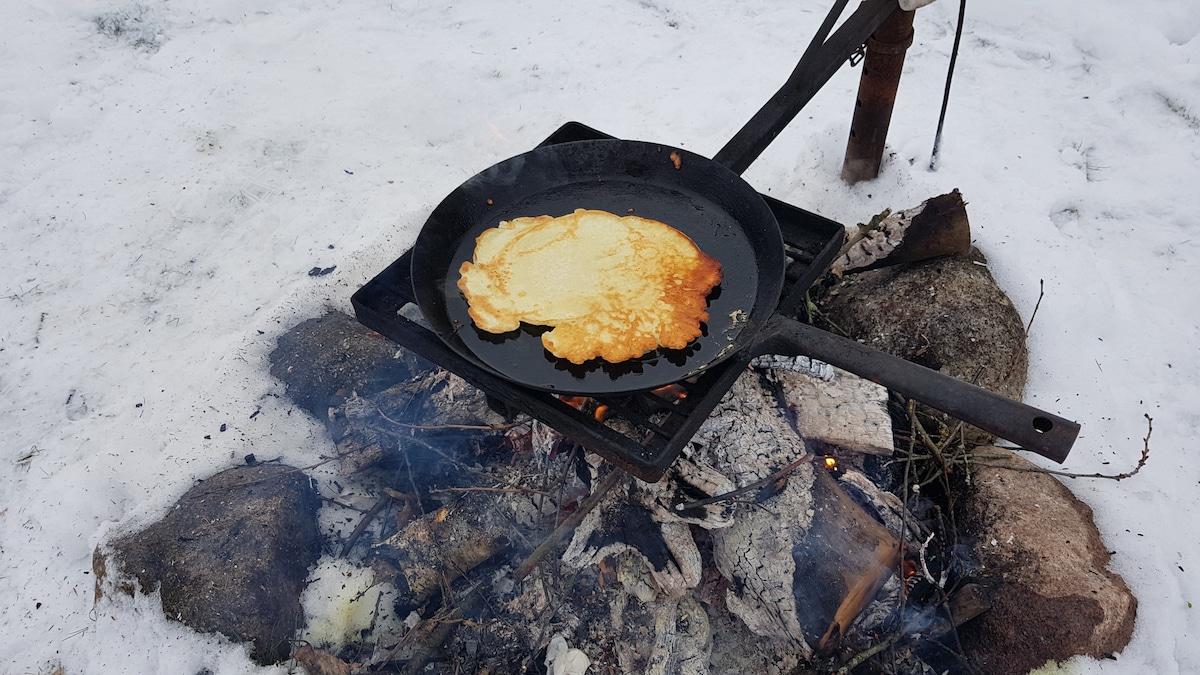 A cast iron skillet is positioned over an open fire, with a pancake cooking on its surface. Snow covers the ground, and glowing embers and smoke are visible around the fire, creating a warm cooking environment in a winter setting.
