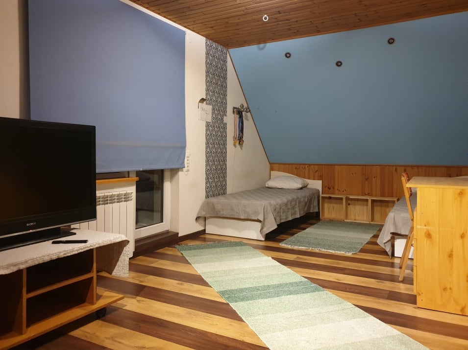 A spacious room features a light blue accent wall and wooden paneling. Two single beds are positioned alongside a simple wooden table. A TV is set on a stand, and a small area rug adds comfort to the wooden floor. Natural light enters through a window with a light blue shade.
