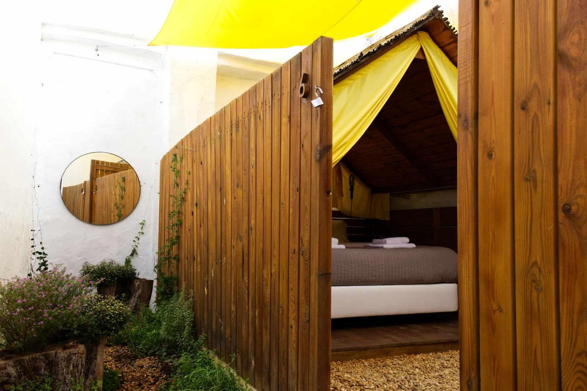 A wooden fence encloses a private outdoor space, with a circular mirror mounted on the wall. Inside, a spacious bed is visible, partially obscured by a yellow canopy and framed by natural greenery surrounding the area.