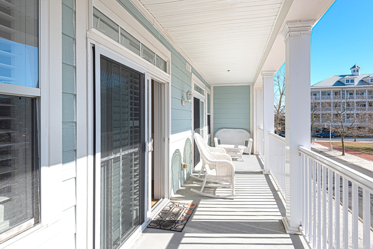 A covered balcony is presented with white wicker furniture, including a sofa and two chairs. Natural light filters through glass doors, illuminating the neutral color scheme of the space. The railing provides a view of the surrounding area, enhancing the inviting outdoor setting.