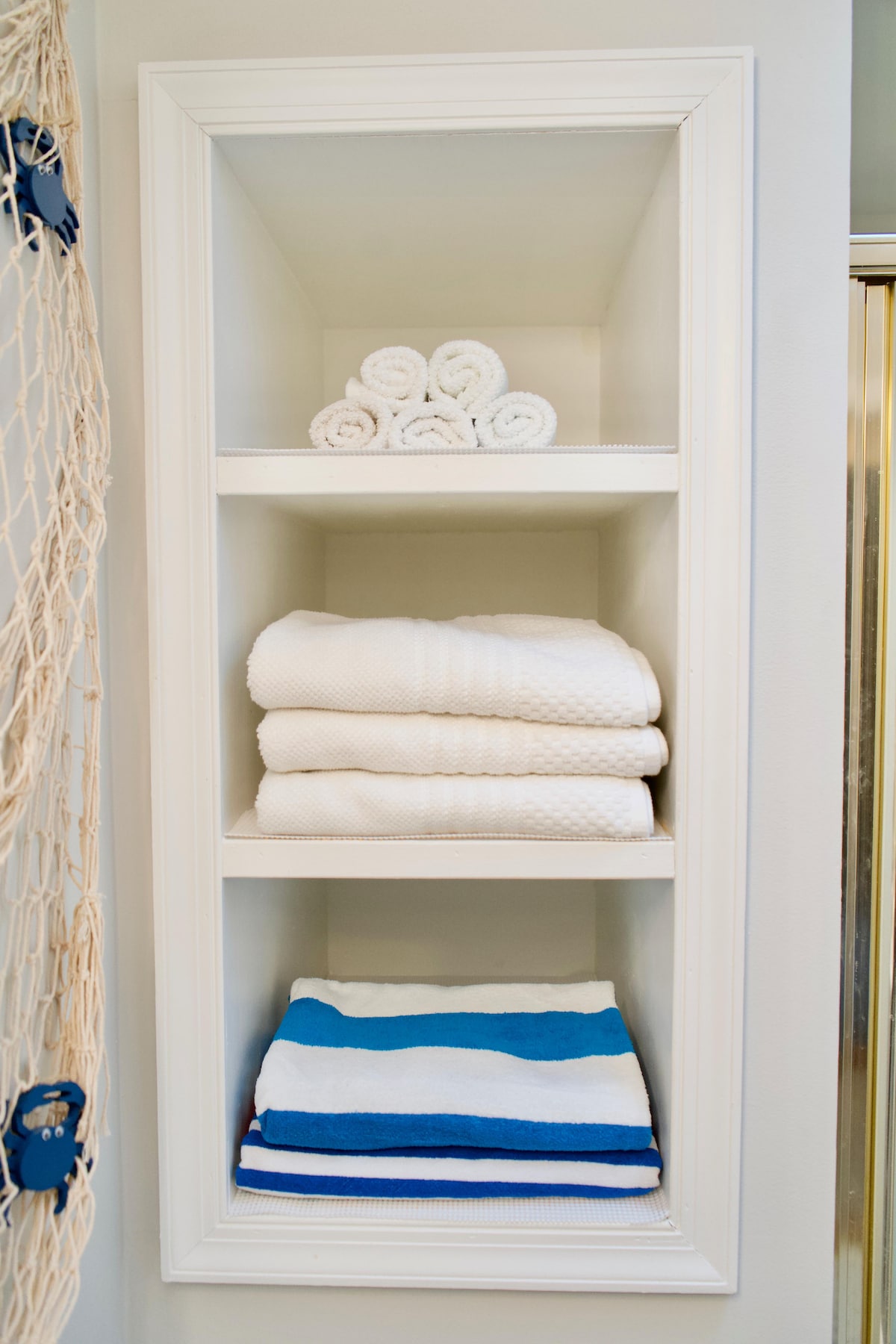 A wall-mounted storage unit features neatly arranged towels. Fresh, white bath towels are stacked on two shelves, while folded beach towels with blue and white stripes occupy the bottom shelf. The overall presentation is clean and organized, suitable for guests' convenience.
