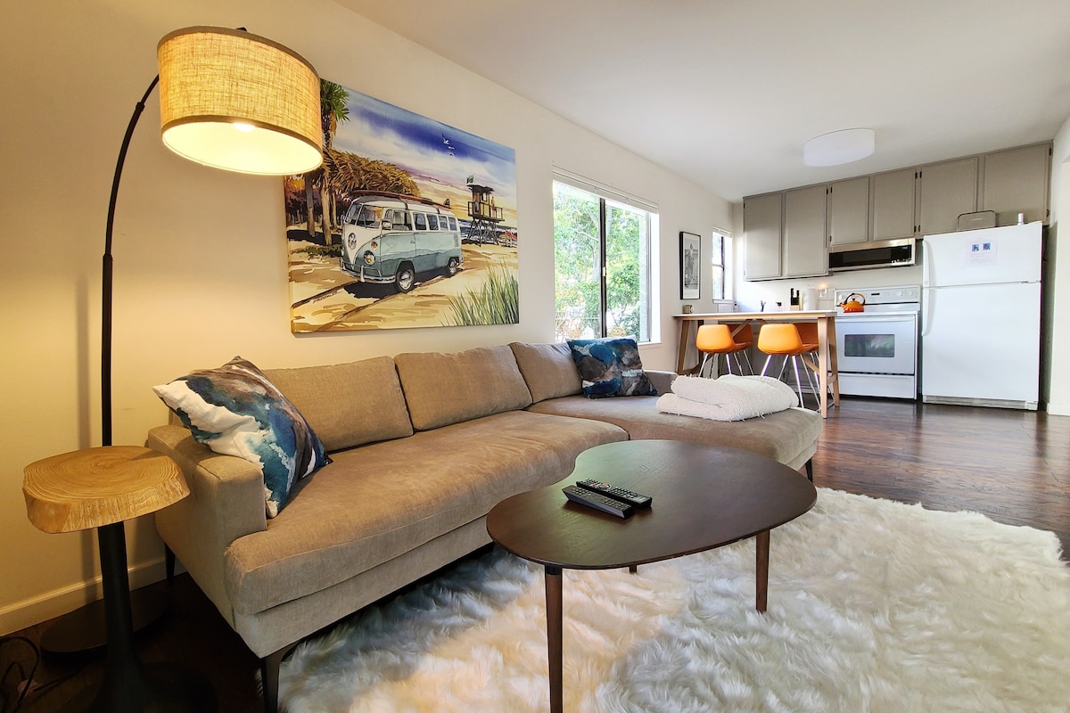A spacious living area features a cozy sectional sofa adorned with decorative pillows. A wooden coffee table sits atop a soft area rug. Natural light pours in through large windows, illuminating the vibrant artwork of a beach scene. The kitchen, equipped with modern appliances, is visible in the background.