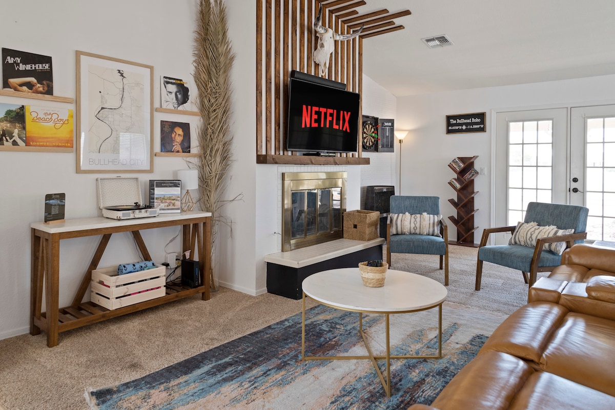A welcoming living room is designed with cozy seating arrangements, including two upholstered chairs and a leather couch. A coffee table rests on a patterned area rug, and a television mounted above the fireplace displays a Netflix logo. Artwork and decorations enrich the wall space.