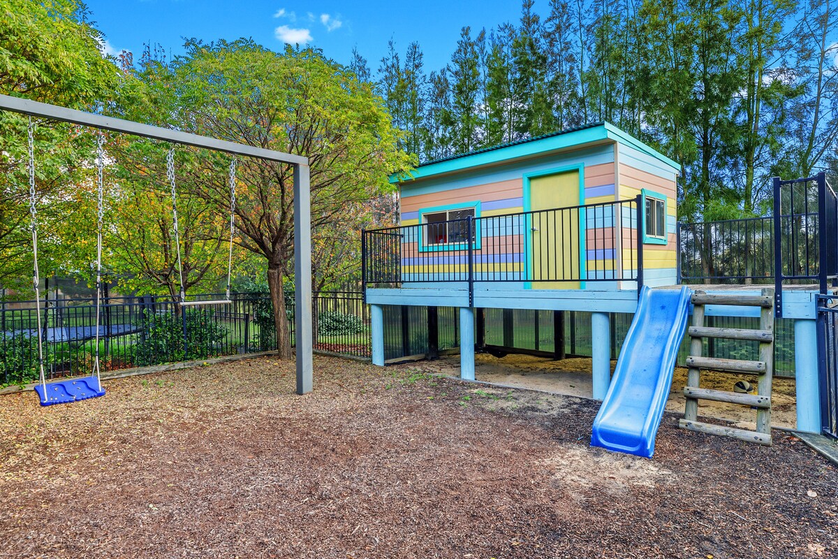 A colorful playground is showcased, featuring a raised cubby house with a vibrant exterior. A blue slide descends from the structure, and swings are positioned nearby. The area is shaded by trees, with soft ground materials for safety.